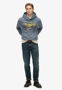 Sudadera azul claro con un estampado gráfico de flores y texto, superpuesta a una camiseta beige, combinada con jeans de mezclilla oscura y zapatos negros.