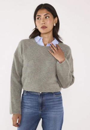 Femme portant un pull gris clair par-dessus une chemise bleue à col et un jean bleu, regardant sur le côté avec une main près du col.