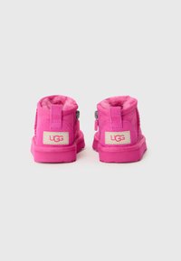 Botas de tobillo de ante rosa con forro de piel de primera calidad, cierre de cremallera y lengüetas en el talón. Presentan una etiqueta rectangular de UGG en crema en la parte posterior.