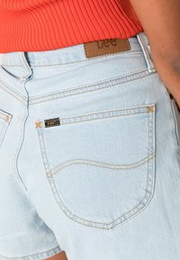 Pantalones cortos de mezclilla de color azul claro con cintura alta. Presenta una etiqueta de cuero marrón y detalles de costura en los bolsillos. Textura suave con un ajuste casual.