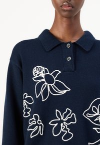 Temno modra pletena polo majica z belim cvetličnim vezenjem, z ovratnikom in dvojnim zapenjanjem. Strukturna površina tkanine.