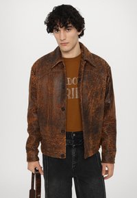 Giacca in pelle marrone con una finitura testurizzata, caratterizzata da un colletto classico, bottoni frontali e due tasche sul petto, abbinata a una camicia marrone.