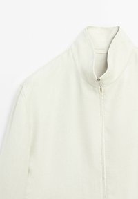 Giacca zip-up di un beige chiaro, con colletto alto, tessuto testurizzato e linee pulite. Caratterizzata da un design minimalista e cuciture sottili.