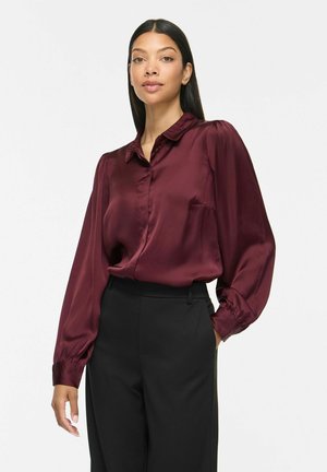Femme aux longs cheveux foncés portant un chemisier en satin bordeaux et un pantalon noir taille haute, debout devant un fond blanc uni.