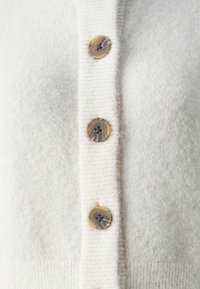 Light beige cardigan made of soft, fluffy material featuring four round, marbled buttons along the front. Vertical ribbing at the hem.