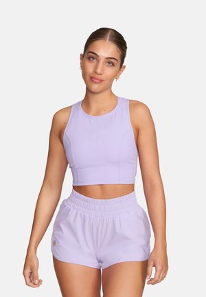 Kobieta z włosami związanymi do tyłu, ubrana w jasnofioletowy, bezrękawnikowy krótki top i pasujące do niego krótkie sportowe szorty z wysokim stanem, stojąca na tle jednolitego tła.