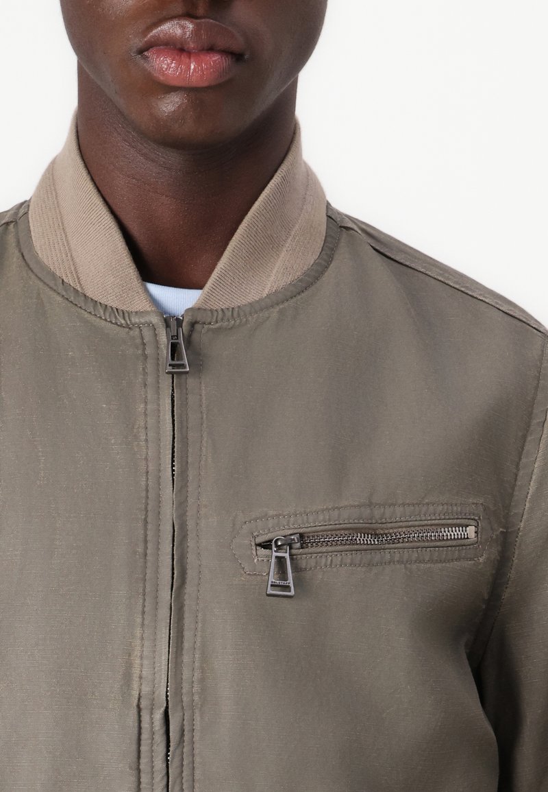 Giubbotto bomber in tessuto verde oliva, con colletto a coste beige e dettaglio della tasca con zip sul lato sinistro del petto. Texture liscia, design casual.