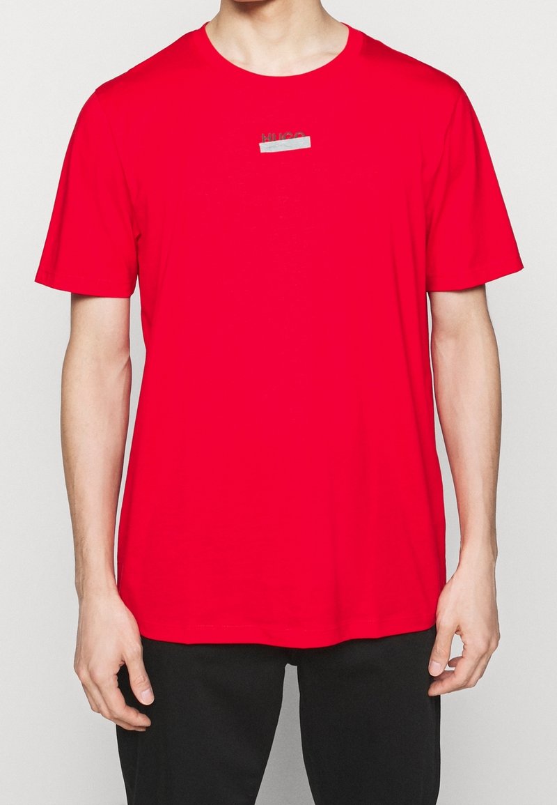 T-shirt rouge à manches courtes en coton, avec un col rond et un petit logo argenté sur la poitrine. Texture lisse.