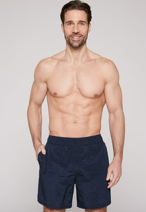 Lächelnder muskulöser Mann mit kurzen braunen Haaren, der marineblaue Badeshorts trägt und mit den Händen in den Taschen vor einem schlichten hellen Hintergrund steht.