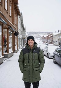 Zielona kurtka puchowa z czarnym pikowanym karczkiem, z zamykanymi na zamek kieszeniami, noszona wraz z czarną dzianinową czapką, na zaśnieżonej ulicy miejskiej.