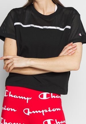 Frau trägt schwarzes Champion Mesh-Kurzarmshirt und rote Leggings mit weißem Champion-Logo-Muster, verschränkte Arme auf der Taille.