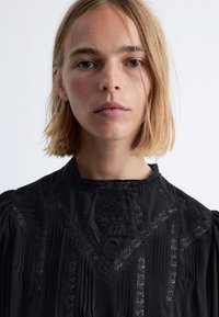 Schwarze Bluse mit Spitzenverzierungen, bestickten Blumenmustern, strukturierten Manschetten und gefältelten Akzenten. Hoher Kragen mit aufwändiger trim.