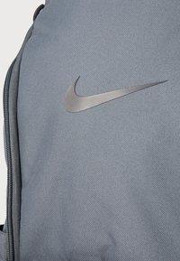 Zaino Nike in tessuto grigio scuro con superficie texturizzata, dotato di un evidente logo Nike swoosh argento. Dettaglio della cerniera visibile.