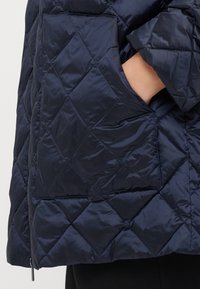 Polstret quilted jakke i mørk marineblå. Har et glat, skinnende stof med diamantformet quiltning og en sidelomme.
