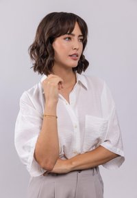 Weißes Hemd mit Knöpfen und einer Tasche, locker getragen. Goldene Armbanduhr mit mehrfarbigen Akzenten. Model mit kurzem, gewelltem Haar.