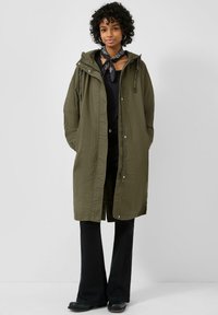 Vrouw die een olijfgroene lange jas met capuchon draagt, een zwarte top, zwarte uitlopende broek, zwarte laarzen en een zwarte sjaal met patroon om haar nek, staand.