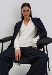 Model trägt ein weißes V-Ausschnitt-Oberteil und dunkelblaue weite Jeans. Eine dunkle Jeansjacke liegt auf ihren Schultern. Sie sitzt auf einem schwarzen Stuhl.
