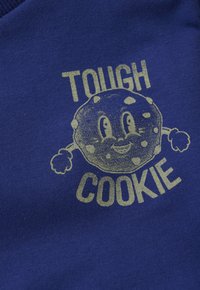 Un biscuit au chocolat souriant en dessin animé avec des bras et des poings, entouré du texte "BISCUIT COSTAUD" sur un tissu bleu.