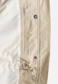 Beige kviltet jakke indeni med en glat hvid foring, udstyret med en sidelomme og justerbar snøre til tilpasning. Sølvknapdetalje.