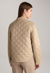 Beige gewatteerde jas met een subtiele glans, voorzien van een platte kraag en een gouden logo op de achterkant, afgewerkt met drukknoopmanchetten.
