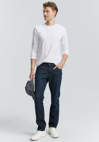 Hvid langærmet skjorte, mørkeblå jeans, holder en grå baseballkasket og parret med hvide sneakers. Simpelt design med en afslappet pasform.