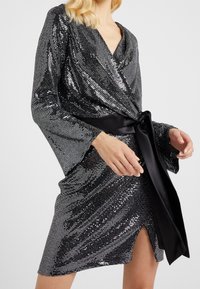 Abito avvolgente nero e argento in paillettes con maniche lunghe, scollatura a V e una cintura in satin legata in vita; presenta uno spacco alto fino alla coscia.