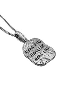 Ciondolo d'argento con una superficie ondulata e texture incisa con "GLI OCCHI REALI RICONOSCONO LE REALI BUGIE", attaccato a una catena metallica.