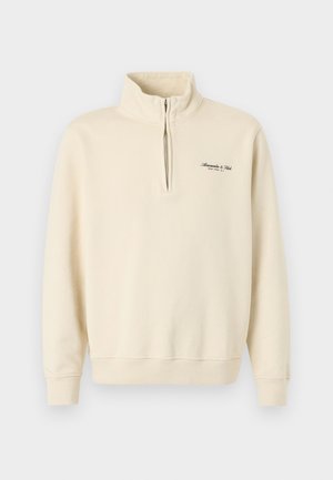 Bēša puszipa džemperis ar augstu apkakli, ribots aproces un apakšmala, kā arī neliels melns logotipa teksts kreisajā krūtīs.