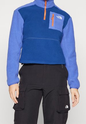 Persoon draagt een blauwe fleece half-ritspullover met een borstzak en zwarte cargobroek, beide met het The North Face-logo.