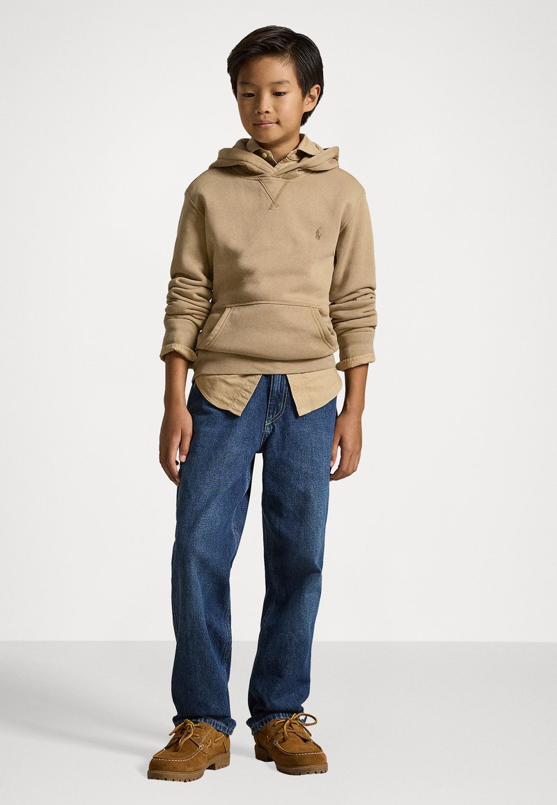 Sweatshirt bege com capuz e bolso frontal, sobre uma camisa clara com colarinho, combinado com jeans de ganga azul e sapatos castanhos.