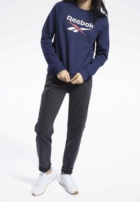 Marinblå sweatshirt med Reebok-logga i rött och vitt, kombinerad med svarta jeans och vita sneakers. Tyget verkar mjukt med en avslappnad passform.