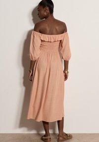 Femme portant une robe midi pêche épaules dénudées avec manches bouffantes, bracelets en or, boucles d’oreilles créoles et sandales marron, debout contre un mur clair.