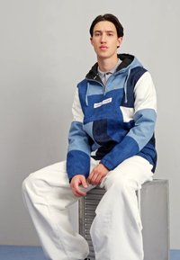 Sininen ja valkoinen tilkkutyöhuppari, jossa on nyörit, yhdistettynä valkoisiin leveisiin housuihin, istumassa metallilaatikolla yksinkertaista taustaa vasten.