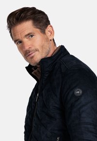 Donkerblauwe gestikte jas met een hoge kraag en rits. Beschikt over een gestructureerde stof en een ronde logo-patch op de linker mouw.