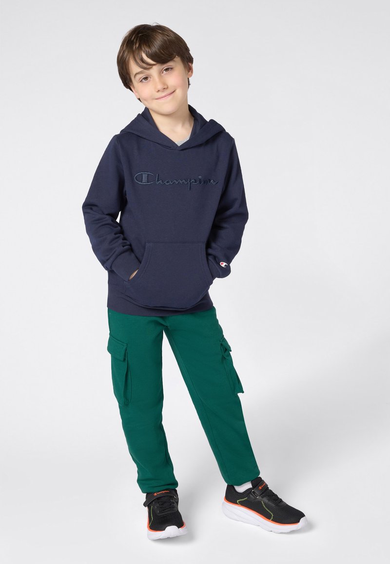 Ragazzo in piedi con le mani nelle tasche, che indossa un felpa Champion blu navy, pantaloni cargo verdi e scarpe nere con dettagli arancioni.