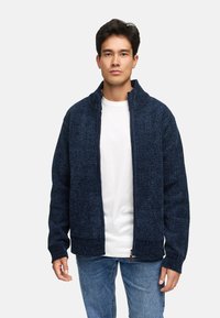 Strikket navy blå zip-jakke med høj krave, ribbede manchetter og en struktureret overflade. Bæres over en hvid skjorte og slim-fit jeans.