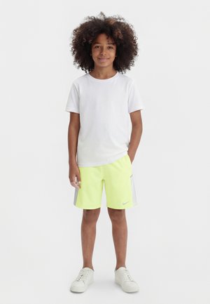 Niño pequeño con cabello rizado de pie sonriendo, vistiendo camiseta blanca, pantalones cortos neón amarillos con rayas grises y zapatillas blancas sobre un fondo liso.