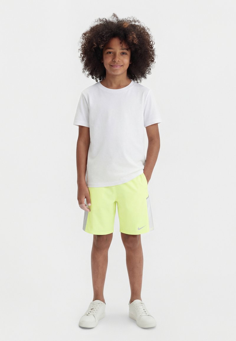 Jeune enfant aux cheveux bouclés, debout et souriant, portant un t-shirt blanc, un short jaune néon avec des rayures grises, et des baskets blanches sur un fond uni.
