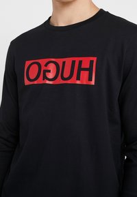 Camiseta negra de manga larga con un logo rectangular rojo brillante en el que se lee la palabra "HUGO" en letras negritas, centrado en el frente.