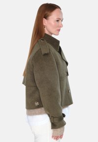 Olivenfarget ulljakke med en avkortet design, bred krage, knappedetaljer og skulderklapper. Båret over en beige genser og hvit bluse.