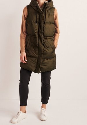 Person trägt olivgrüne lange Steppweste mit Kapuze, beige langärmeliges Oberteil, schwarze Hose und weiße Turnschuhe, steht vor einfarbigem Hintergrund.