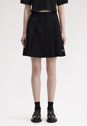 Persona con falda negra plisada con logo lateral, top corto negro, calcetines negros y mocasines negros, de pie frente a un fondo blanco.