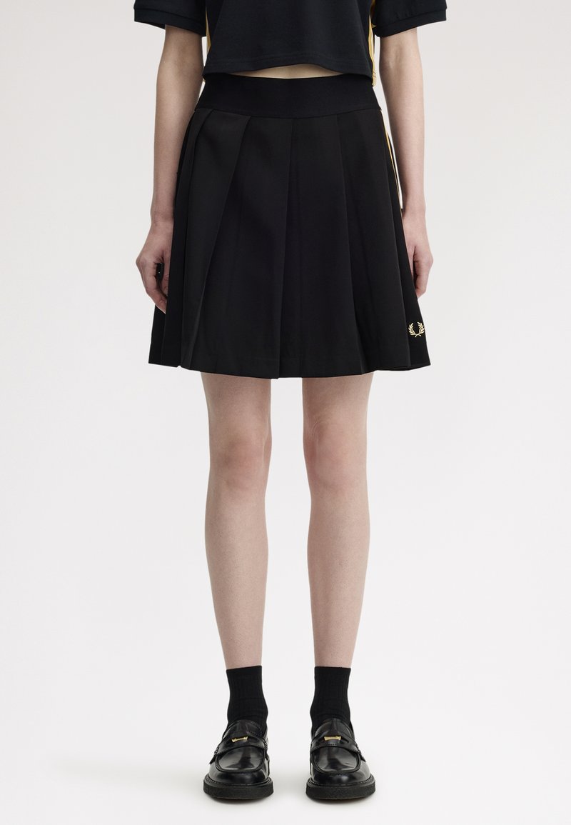 Personne portant une jupe plissée noire avec un logo latéral, un haut court noir, des chaussettes noires et des mocassins noirs, debout devant un fond blanc.