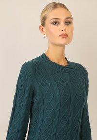 Teal kabelstrikket sweater med ribbet rund halsudskæring, der har strukturerede diamant- og snoede mønstre, lavet af et blødt og tykt materiale.