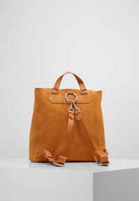 Mochila de ante en amarillo mostaza con forma rectangular, asa superior, correas ajustables de cuero y un detalle metálico plateado.