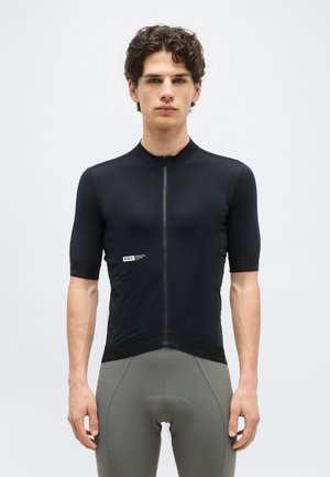 Mladý muž oblečený v priliehavom čiernom cyklistickom drese s krátkymi rukávmi a sivých cyklistických kraťasoch s vypchávkami, stojaci pred jednoduchým pozadím.