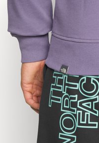 Lila sweatshirt med ribbade ärmslut, med en liten svart etikett. Handen vilar mot svarta byxor med stort turkos tryck.