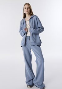 Completo tuta azzurra chiara caratterizzato da una felpa con zip e dettagli in strass e pantaloni della tuta larghi con decorazioni coordinate.