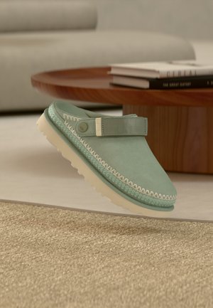 Chaussure à enfiler vert clair en daim avec des coutures blanches et une sangle. Elle possède une semelle en caoutchouc texturée pour l'adhérence et le confort.