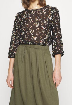Kobieta ubrana w czarną, przezroczystą, kwiatową bluzkę z rękawami do połowy przedramienia, wpuszczoną do wysokiej talii oliwkowozielonej spódnicy.